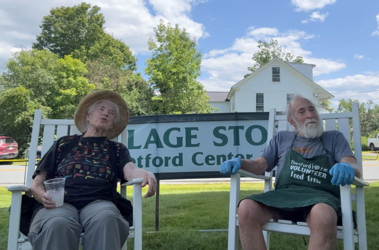Two volunteers relax in white rocking chairs in front of the Village Store sign, one wearing a Thetford Hill Fair Food Crew apron