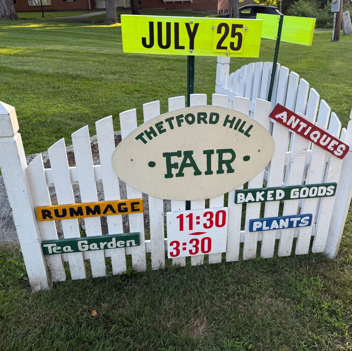 A hand-painted sign on a white picket fence announcing the Thetford Hill Fair on July 25, 11:30 to 3:30, with rummage, tea garden, antiques, baked goods, and plants