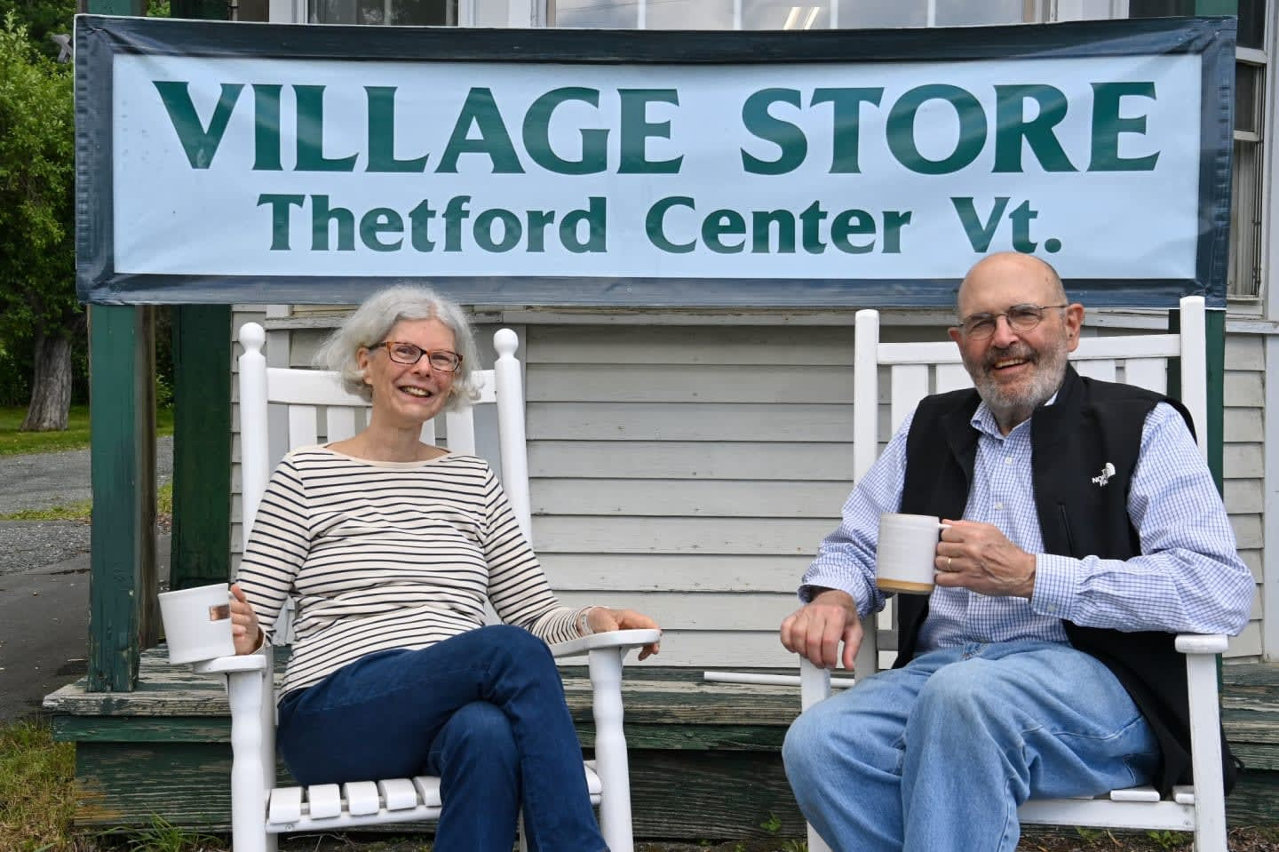 A woman and man relax in white rocking chairs with coffee mugs beneath the Village Store, Thetford Center Vt. sign