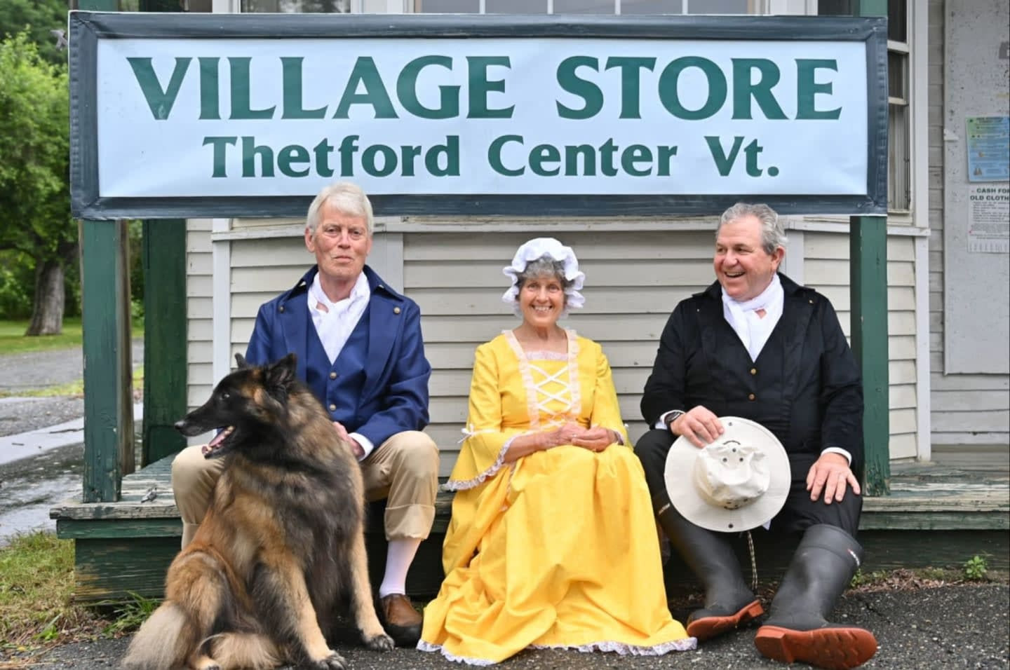 Three community members in colonial-era costume, with a dog, seated beneath the Village Store sign