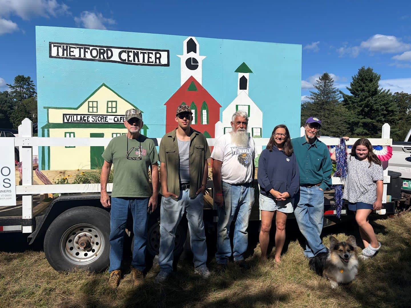 Volunteers and a corgi stand in front of the Thetford Center parade float, a painted mural of the Village Store & Café, Town Hall, and church on a trailer